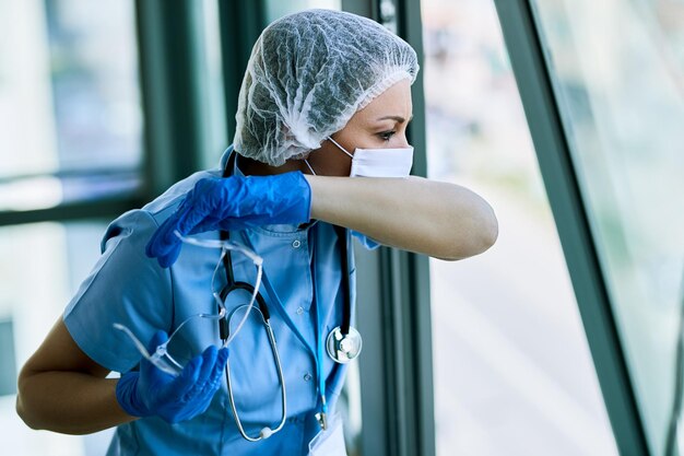 Enfermeira vestindo roupas de trabalho protetoras e espirrando no cotovelo enquanto trabalhava na clínica médica durante a epidemia de coronavírus