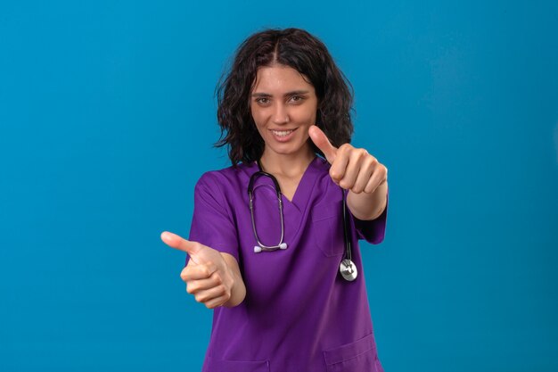 enfermeira vestindo uniforme e estetoscópio com sorriso confiante mostrando os polegares para cima com cara feliz em pé no azul isolado