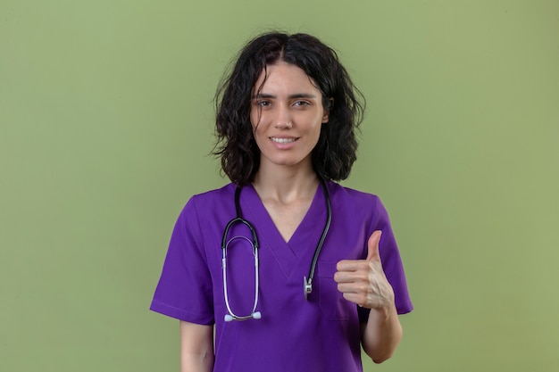 enfermeira vestindo uniforme e estetoscópio com um sorriso confiante mostrando o polegar em pé no verde isolado