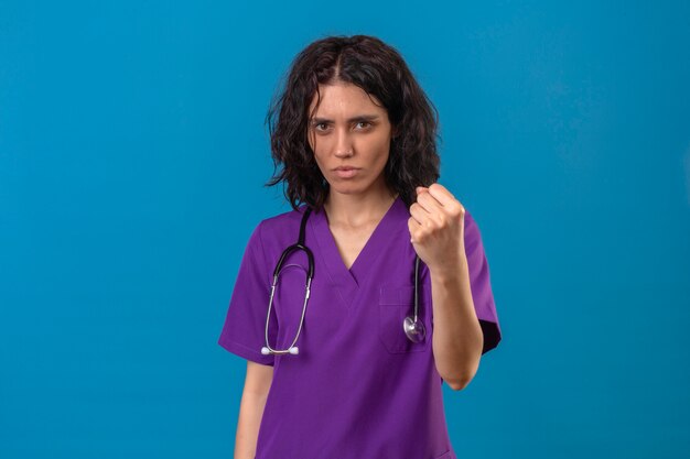 enfermeira vestindo uniforme e estetoscópio mostrando punho para a câmera com rosto carrancudo e ameaçador em azul isolado