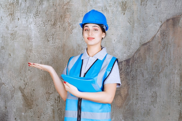 Engenheira de uniforme azul e capacete segurando uma pasta de relatório azul e apontando para alguém ao redor.