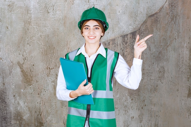 Engenheira de uniforme verde e capacete segurando uma pasta de projeto verde e apontando para o lado direito.
