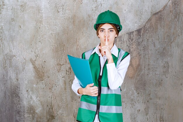 Engenheira de uniforme verde e capacete segurando uma pasta de projeto verde e pedindo silêncio.