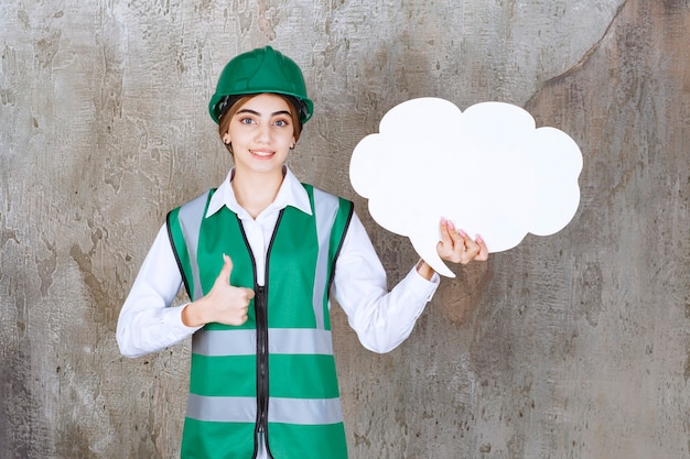 Engenheira de uniforme verde e capacete segurando uma placa de informações em forma de nuvem e mostrando sinal com a mão positiva