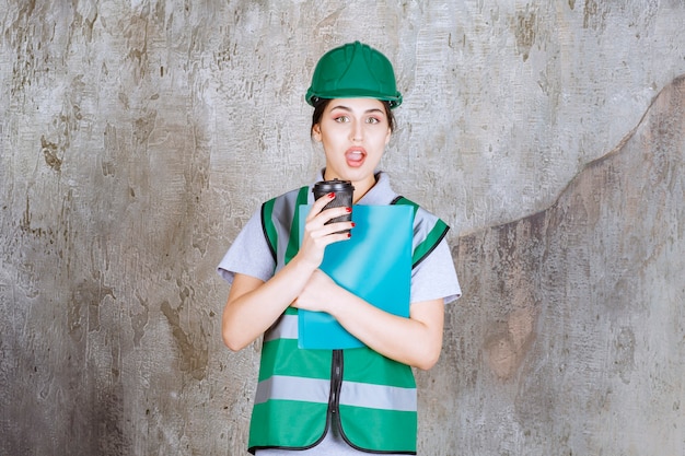 Engenheira de uniforme verde e capacete segurando uma xícara de café preta e uma pasta de projeto azul