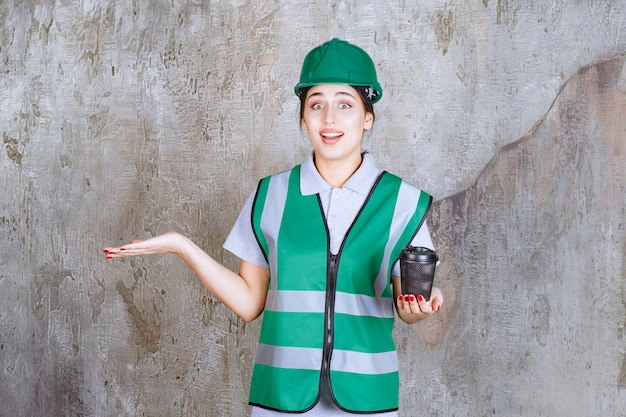 Engenheira de uniforme verde e capacete segurando uma xícara de café preto e mostrando algo à parte.