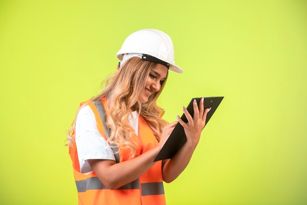 Engenheira em capacete branco e equipamento segurando a lista de verificação e tomando notas.