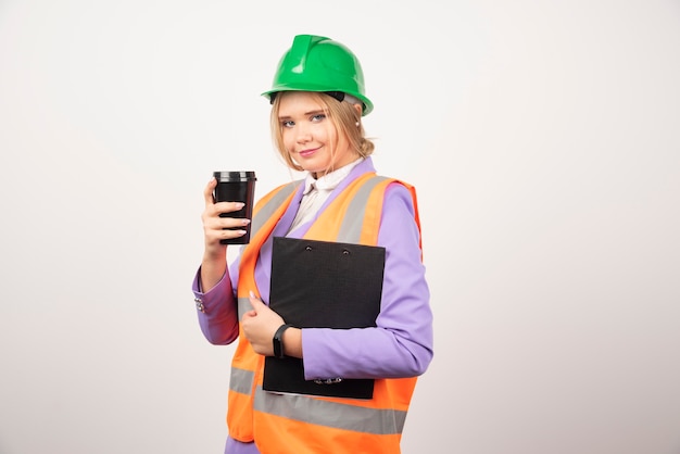 Engenheira industrial feminina de uniforme com área de transferência e copo preto na parede branca.