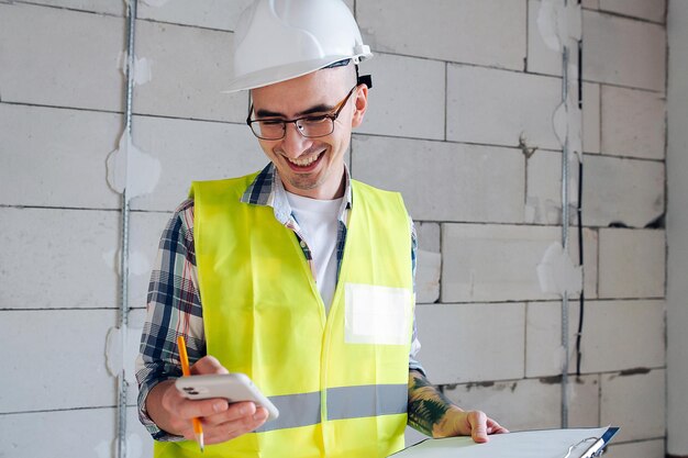 Engenheiro de construção feliz no capacete branco olhando para seu telefone, rolando. sorrindo amplamente. dentro de um prédio inacabado