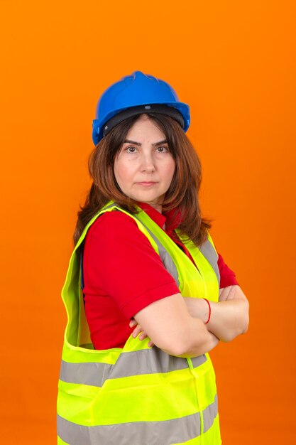 Engenheiro de mulher vestindo colete de construção e capacete de segurança em pé lateralmente com os braços cruzados com cara séria sobre parede laranja isolada