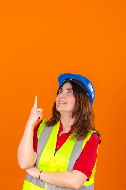 Foto grátis engenheiro de mulher vestindo colete de construção e capacete de segurança olhando para cima apontando para algo com sorriso no rosto sobre parede laranja isolada