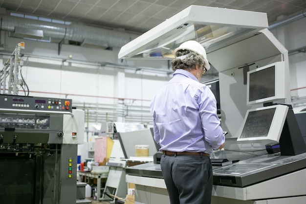 Engenheiro de planta masculino de cabelos grisalhos, capacete e óculos, parado na máquina industrial, usando dispositivo digital