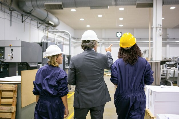 Engenheiro e funcionárias de fábrica em capacetes, andando no chão da fábrica e conversando, homem apontando para o equipamento e instruindo mulheres