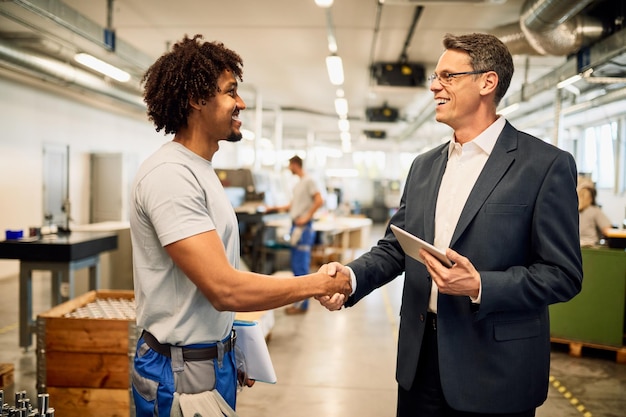 Engenheiro feliz e jovem trabalhador negro apertando as mãos enquanto cumprimenta no edifício industrial