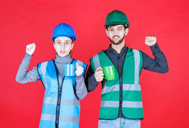 Engenheiros masculinos e femininos com capacete segurando canecas azuis e verdes e mostrando sinal de satisfação.