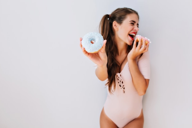 Engraçada jovem bonita com cabelo comprido em body rosa se divertindo com donuts coloridos. jovem alegre com corpo esguio, comendo doces. divertido, estilo de vida brilhante, dieta.