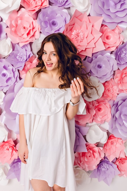 Engraçada linda garota morena com um vestido branco fora do ombro está em um estúdio com fundo de flores rosa.