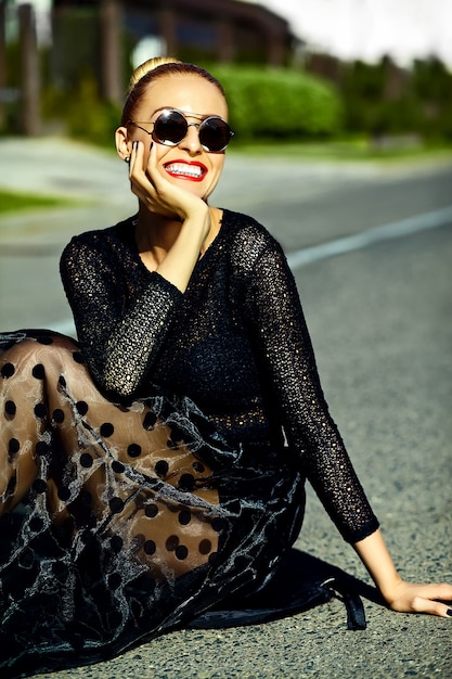 Engraçado elegante sexy sorridente modelo mulher jovem e bonita loira em roupas de verão preto hipster sentado na rua