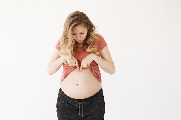 Engraçado Europeu jovem grávida bonita com cabelos longos claros em roupas confortáveis, olhando para a barriga com expressão interessada.