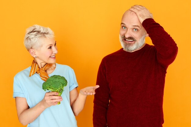 Engraçado pensionista barbudo segurando a mão em seu peito com olhar confuso e intrigado, não quer comer brócolis que a esposa lhe ofereceu