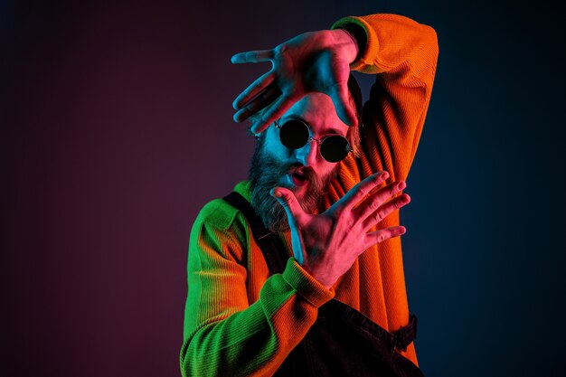 Enquadramento, selfie. Retrato do homem caucasiano em fundo gradiente de estúdio em luz de néon. Lindo modelo masculino com estilo hippie. Conceito de emoções humanas, expressão facial, vendas, anúncio.