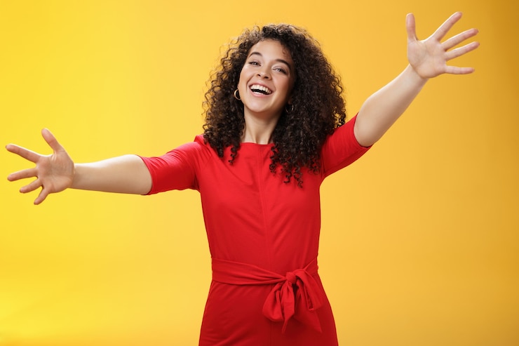 Foto grátis entre em meus braços. retrato de mulher encantadora amigável e amorosa, carinhosa com cabelo encaracolado em vestido vermelho casual espalhar as mãos como querendo dar um abraço, sorrindo amplamente para a câmera dando boas-vindas calorosas ou carinho.