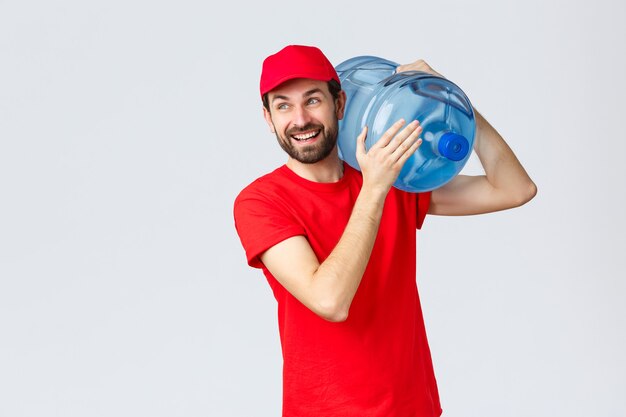 Entrega de comida e mantimentos para viagem, conceito de pedidos sem contato covid-19. Correio barbudo sorridente alegre em uniforme vermelho, boné, traga água engarrafada para o escritório ou casa, desvie o olhar alegre