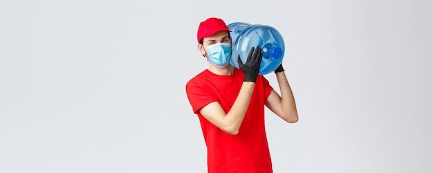 Entrega de comida e mantimentos para viagem conceito de pedidos sem contato covid19 Correio jovem com boné uniforme vermelho e máscara facial com luvas entregando água engarrafada em seu escritório ou casa