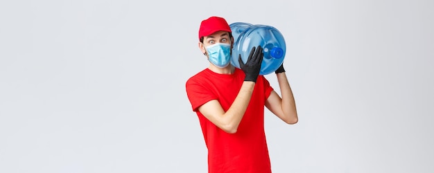 Entrega de comida e mantimentos para viagem conceito de pedidos sem contato covid19 Correio surpreso em luvas de boné uniforme vermelho e máscara facial olhando chocado ao trazer água engarrafada para o escritório ou para casa