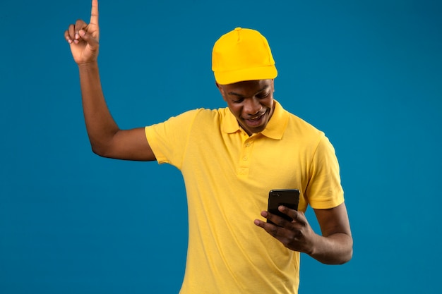 Entregador afro-americano com camisa pólo amarela e boné olhando para a tela do celular apontando o dedo para cima sorrindo em pé no azul