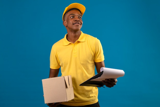 Entregador afro-americano com uma camisa pólo amarela e boné segurando a prancheta e uma caixa de papelão com um sorriso confiante parado no azul isolado