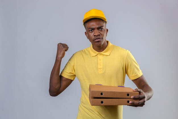 Entregador afro-americano de camisa pólo amarela e boné segurando caixas de pizza levantando o punho com expressão de raiva ameaçando ficar de pé