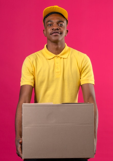Entregador afro-americano de camisa pólo amarela e boné segurando uma caixa de papelão, parecendo confiante em pé na rosa