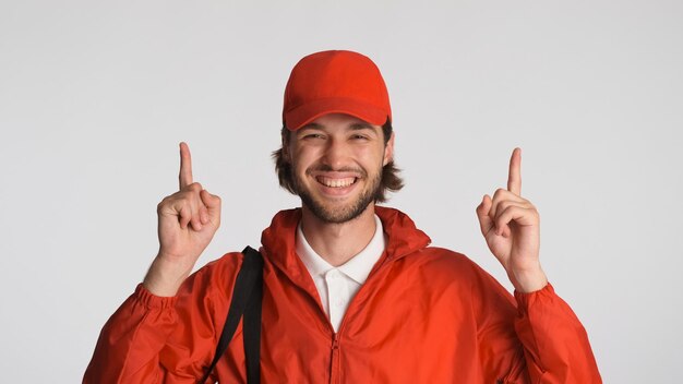 Entregador alegre vestido de uniforme vermelho apontando o dedo indicador para cima no espaço para propaganda ou promoção sobre fundo branco