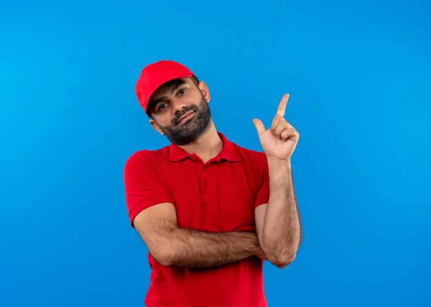 Entregador barbudo com uniforme vermelho e boné apontando com o dedo para o lado, parecendo confiante em pé sobre a parede azul