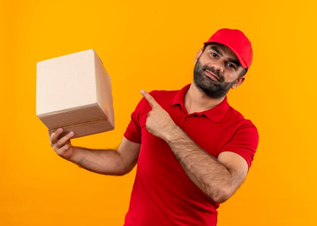 Entregador barbudo com uniforme vermelho e boné segurando a embalagem da caixa apontando com o dedo para ela sorrindo confiante em pé sobre a parede laranja
