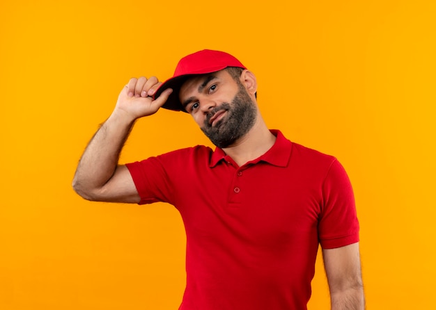 Entregador barbudo de uniforme vermelho e boné com um sorriso no rosto tocando seu boné em pé sobre a parede laranja