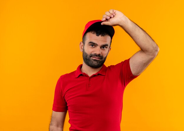 Entregador barbudo de uniforme vermelho e boné com um sorriso no rosto tocando seu boné em pé sobre a parede laranja