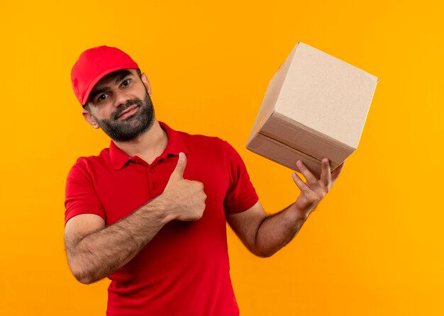 Entregador barbudo de uniforme vermelho e boné segurando a embalagem da caixa mostrando o polegar para cima, positivo e feliz em pé sobre a parede laranja