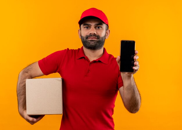 Entregador barbudo de uniforme vermelho e boné segurando a embalagem da caixa mostrando o smartphone parecendo confiante em pé sobre a parede laranja