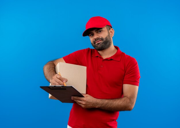 Entregador barbudo de uniforme vermelho e boné segurando o pacote da caixa e a prancheta sorrindo confiante em pé sobre a parede azul