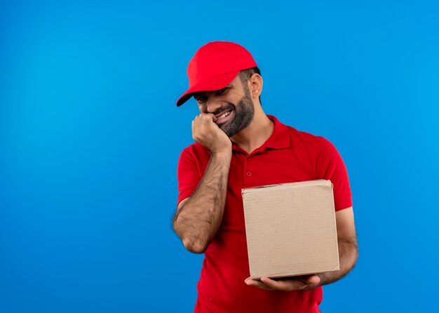 Entregador barbudo de uniforme vermelho e boné segurando um pacote de caixa sorrindo feliz e positivo em pé sobre a parede azul