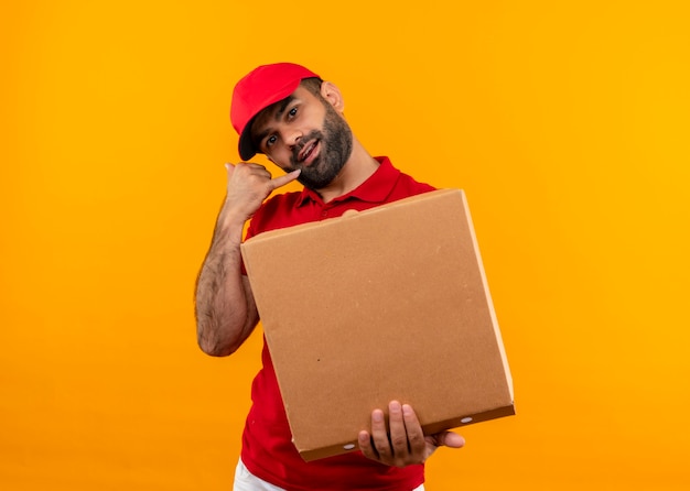 Entregador barbudo de uniforme vermelho e boné segurando uma caixa de pizza aberta, fazendo gesto de me ligar em pé sobre a parede laranja