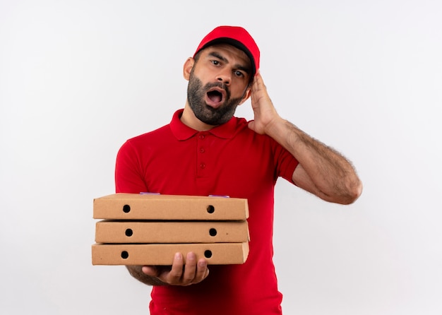 Entregador barbudo de uniforme vermelho e boné segurando uma pilha de caixas de pizza chocado e confuso com a boca escancarada de pé sobre uma parede branca