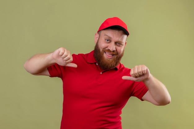 Entregador barbudo jovem satisfeito de uniforme vermelho e boné apontando para si mesmo, sorrindo confiante