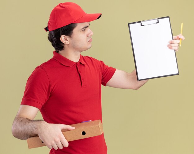 Entregador caucasiano jovem confuso de uniforme azul e boné segurando um pacote de pizza e uma prancheta com um lápis olhando para a prancheta