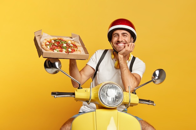 Foto grátis entregador de aparência amigável com capacete dirigindo uma scooter amarela enquanto segura uma caixa de pizza