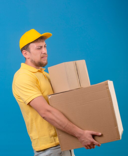 entregador de camisa pólo amarela e boné em pé com grandes caixas de papelão sofrendo com o peso pesado no azul