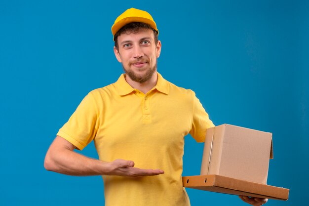 entregador de camisa pólo amarela e boné segurando caixas de papelão, sorrindo amigavelmente e apresentando-se com a palma da mão em pé no azul