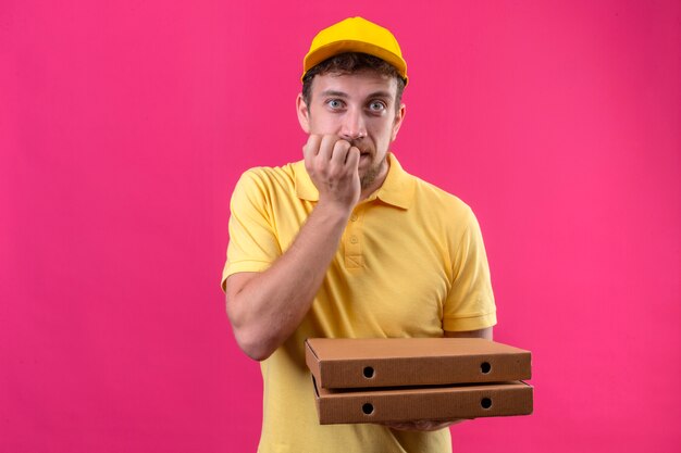 entregador de camisa pólo amarela e boné segurando caixas de pizza, parecendo preocupado, nervoso roendo as unhas em pé no rosa isolado